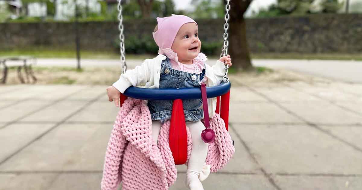 Dieťa s korunkovou čelenkou a pletenou dekou na ihrisku, štýlový doplnok pre bábätká vhodný na prechádzky aj fotenie.