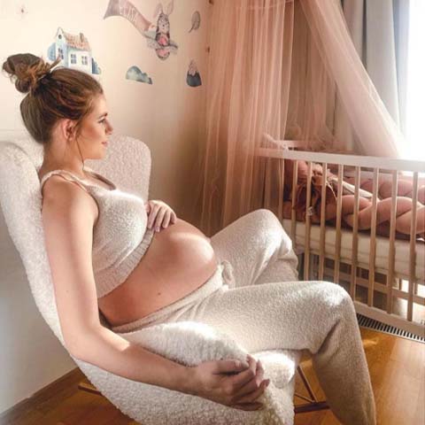 Tehotná žena sediaca v kresle pri detskej postieľke a premýšľa čo ešte potrebuje pre bábätko